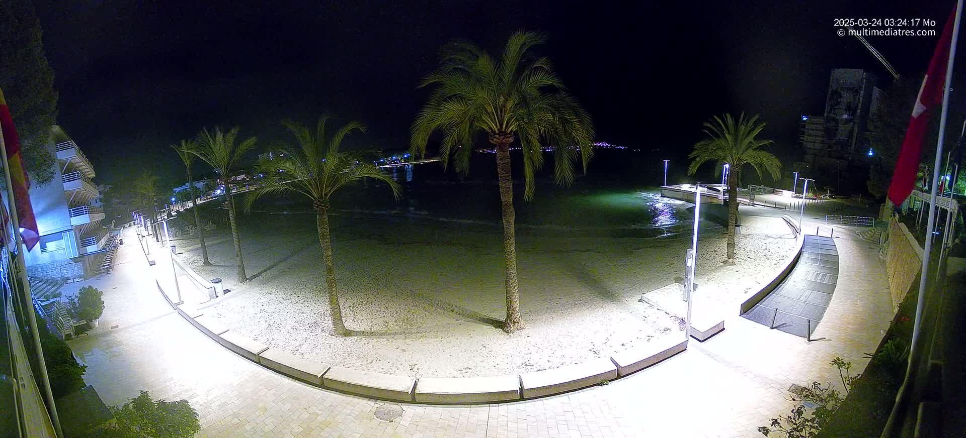 A nighttime, illuminated view of a sandy beach bordered by a paved walkway, palm trees, and buildings, with calm water visible in the background.