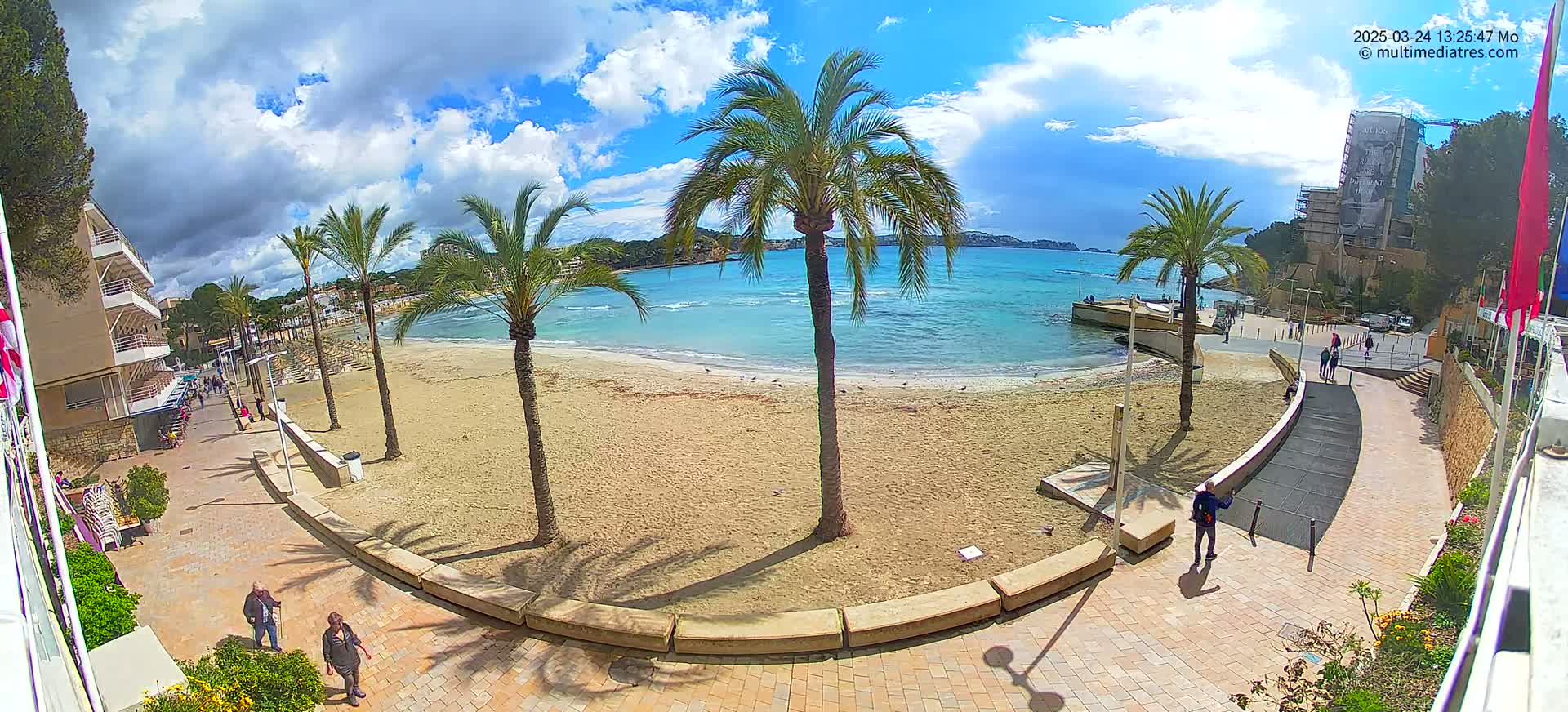 A mostly sunny day at a sandy beach with palm trees, buildings, and people walking along a paved path.