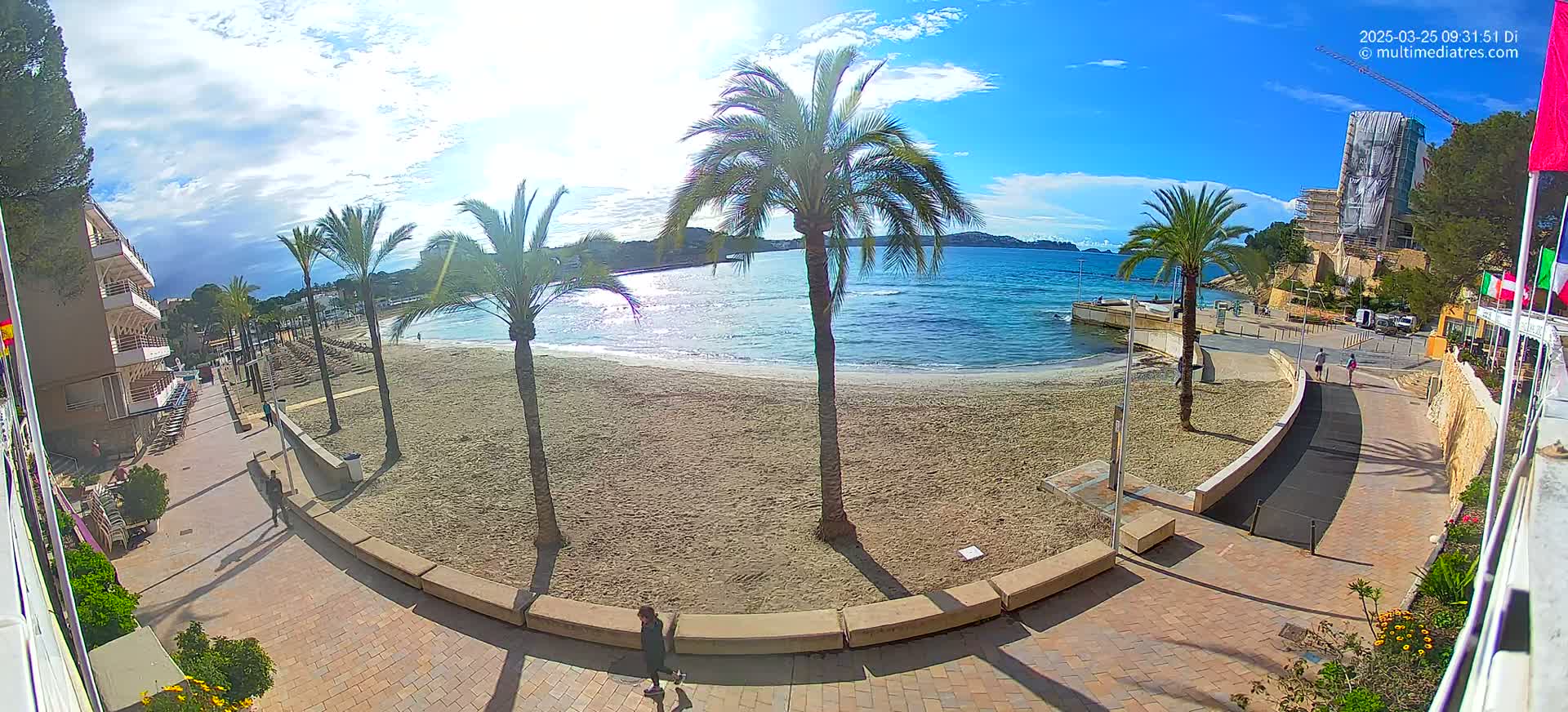A sandy beach with palm trees and a paved walkway leading to buildings is visible under a mostly sunny sky.