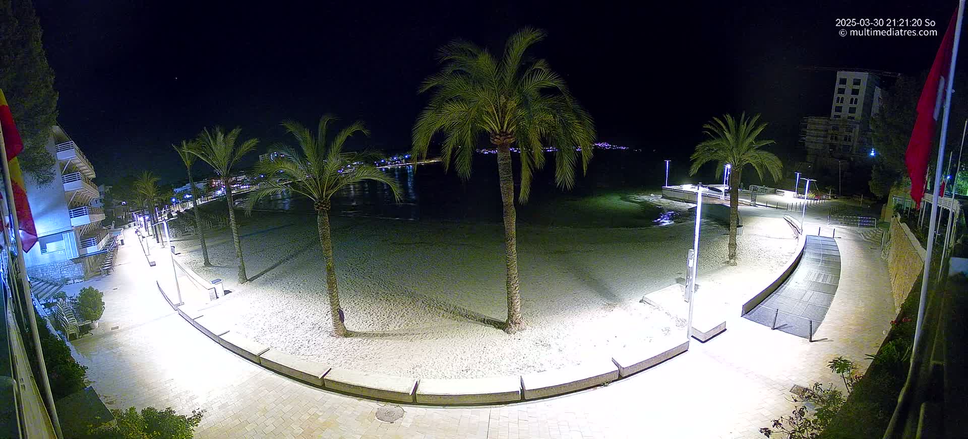 A nighttime view of a sandy beach bordered by a paved walkway, palm trees, and buildings, illuminated by artificial light.
