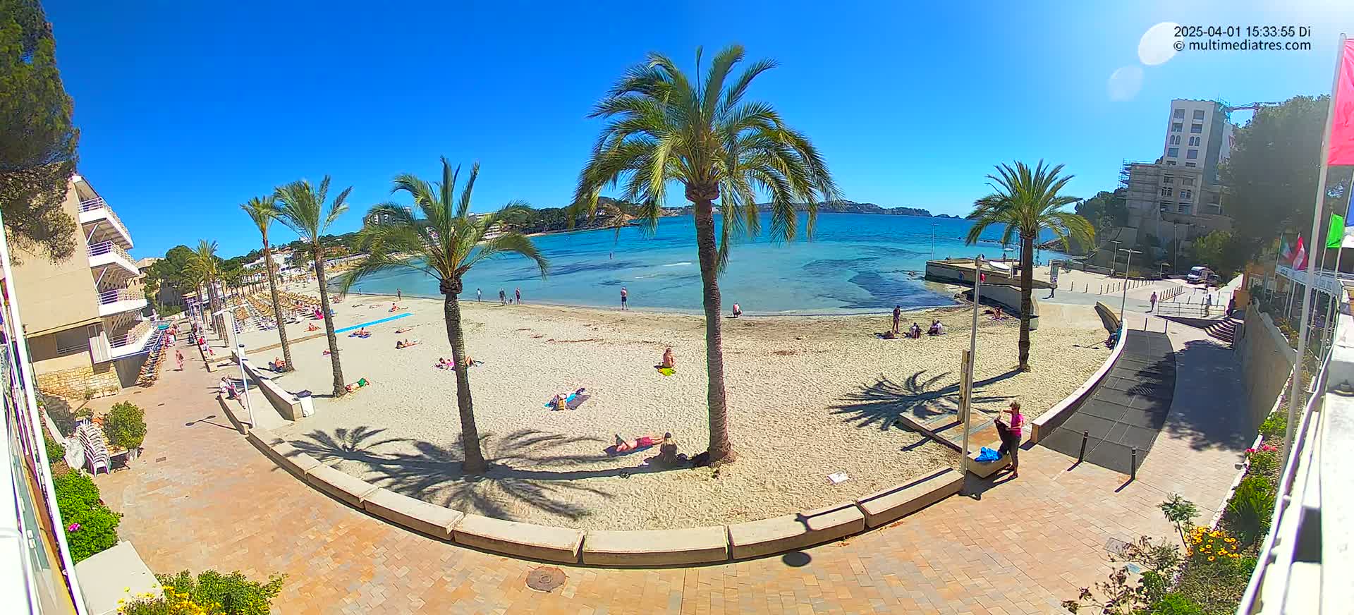 A sandy beach with palm trees and sunbathers is viewed from above on a clear, sunny day, adjacent to buildings and a paved walkway.