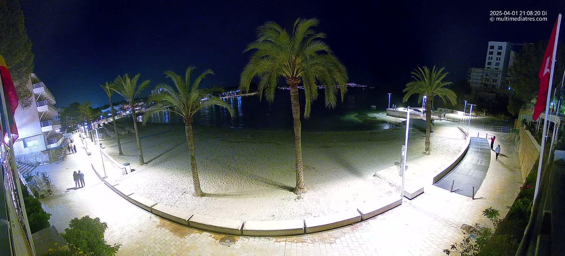 A nighttime view of a sandy beach illuminated by lights, with palm trees lining a paved walkway next to a calm body of water.
