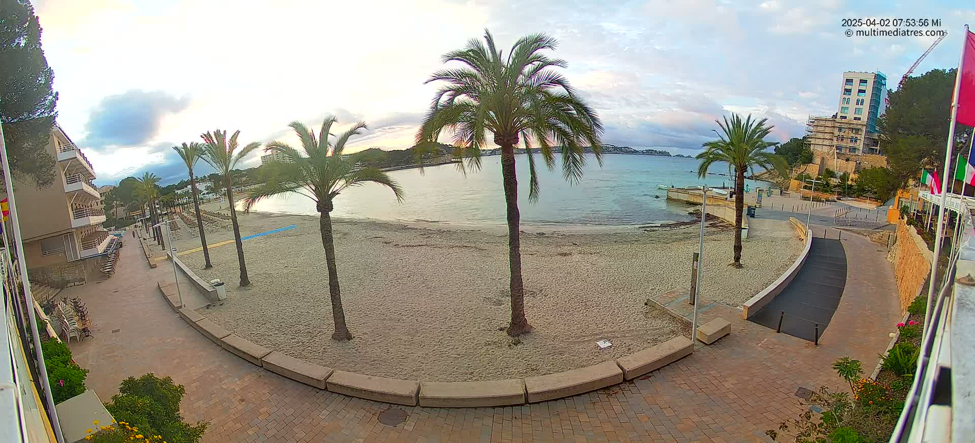 A sandy beach with palm trees and a paved walkway is seen on a partly cloudy morning, overlooking a calm body of water with buildings in the background.