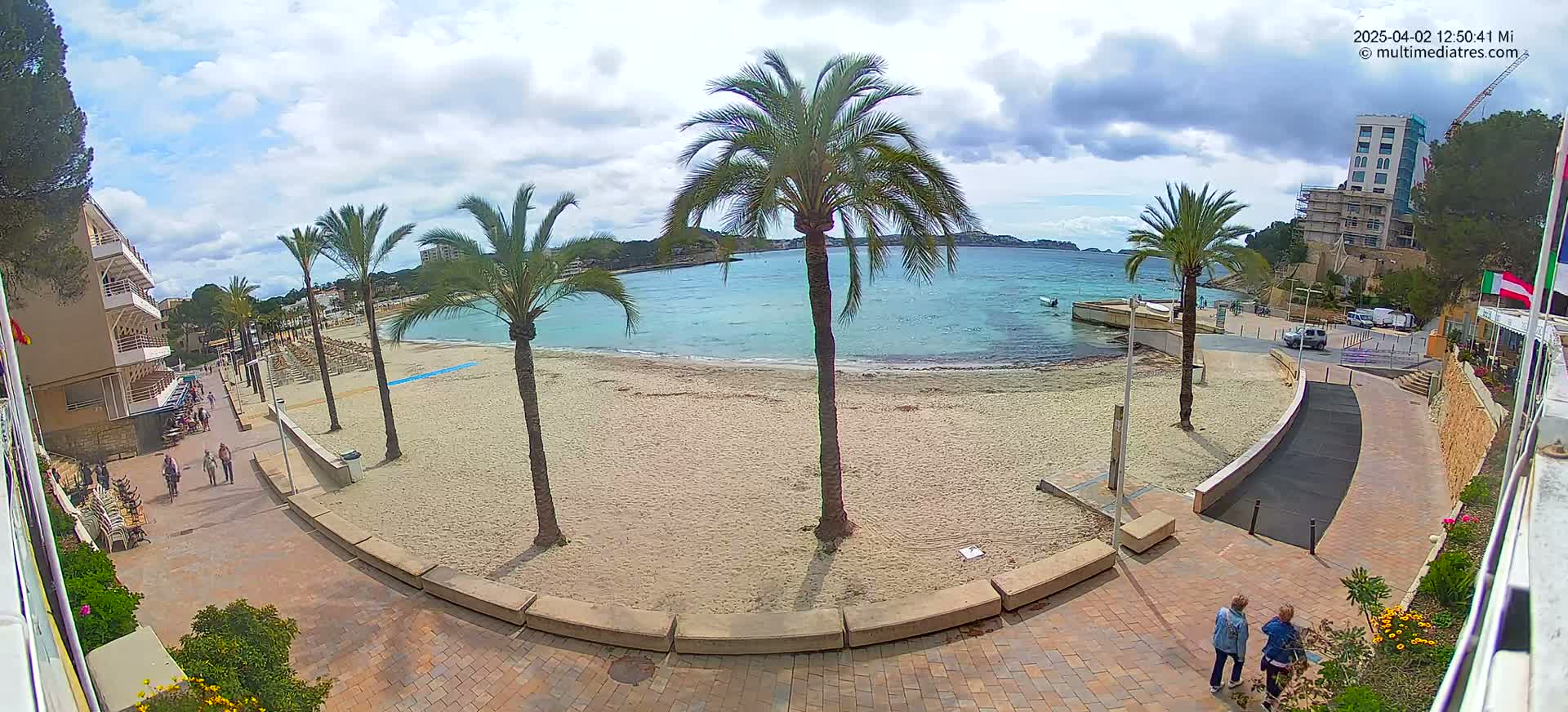 A sandy beach with calm, light-blue water is seen from above on a partly cloudy day, with palm trees lining the shore and people walking on a paved path.