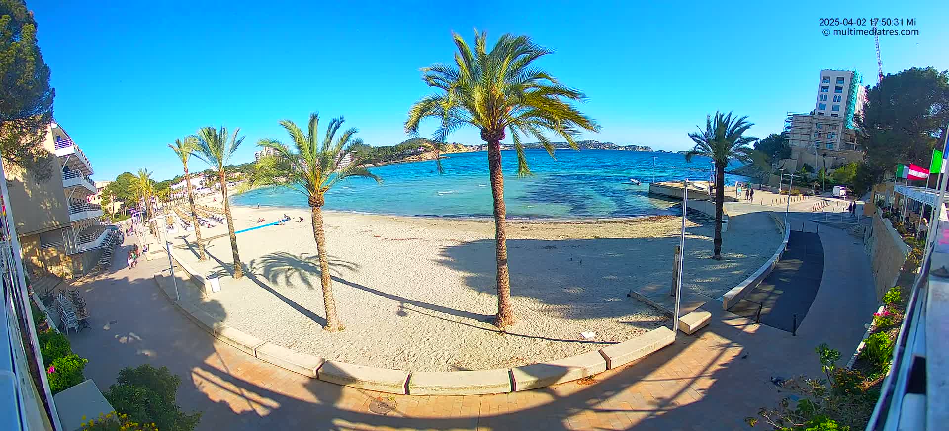 A sandy beach with palm trees and turquoise water under a clear blue sky is viewed from above a paved walkway.