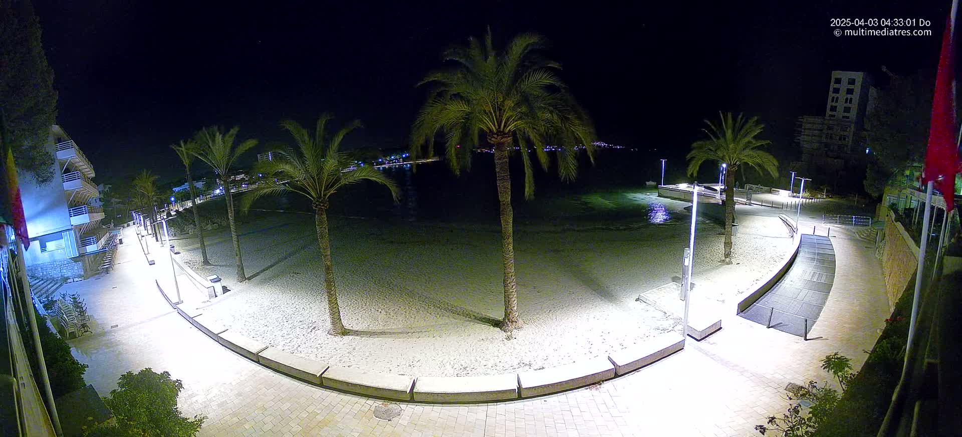A nighttime aerial view of a sandy beach, palm trees, and a paved walkway illuminated by lights, overlooking calm water.