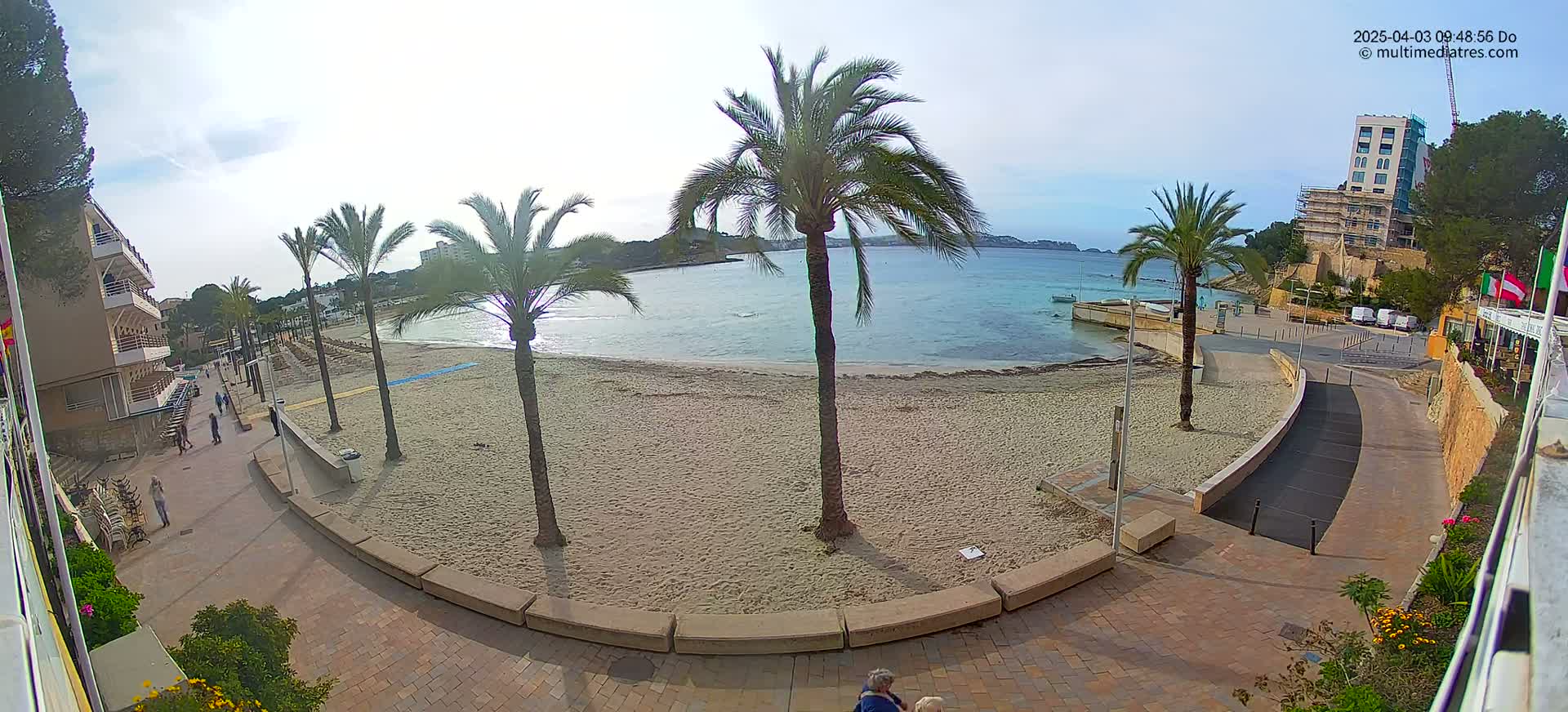 A sandy beach with calm, light blue water is bordered by a paved walkway, palm trees, and buildings under a mostly clear sky.