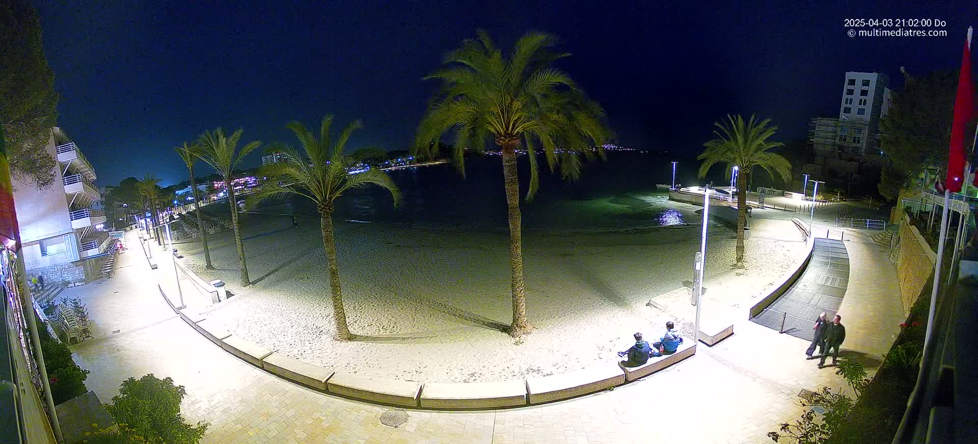 A nighttime view of a sandy beach with palm trees, a paved walkway, and a few people sitting on a bench, under a clear, dark sky.