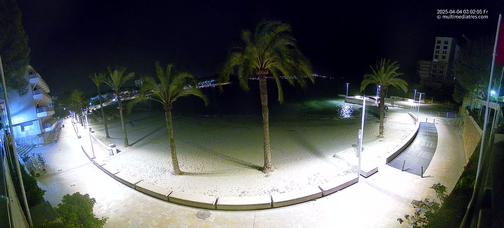 A nighttime, illuminated view of a sandy beach with palm trees, a paved walkway, and buildings in the background.