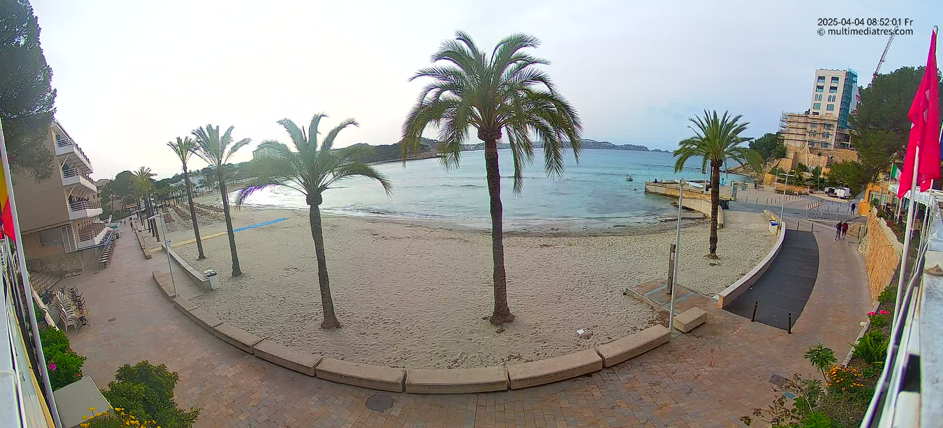 A sandy beach with palm trees and a paved walkway leading to buildings, under a mostly cloudy sky.
