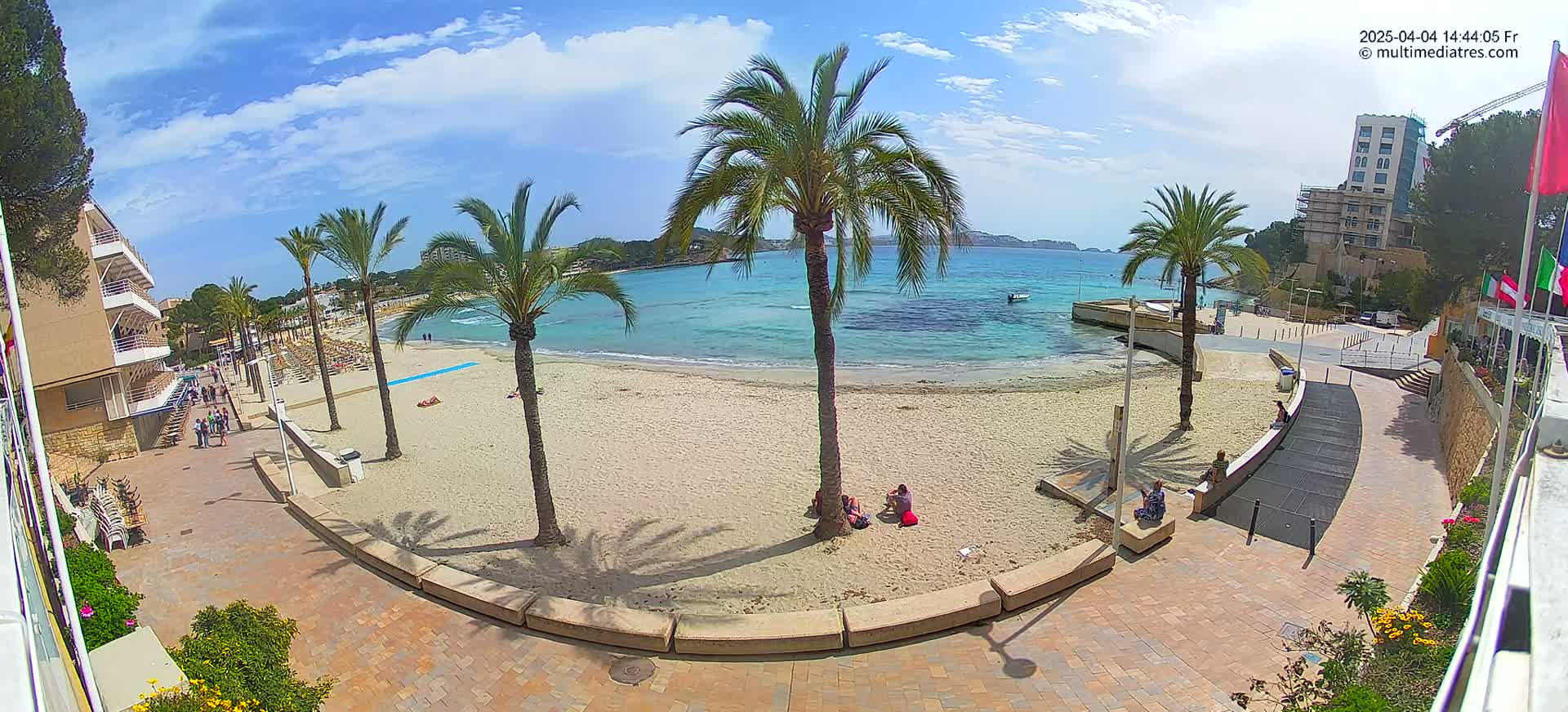 A sandy beach with palm trees and a few people on it is situated next to a paved walkway and buildings, under a partly cloudy sky.