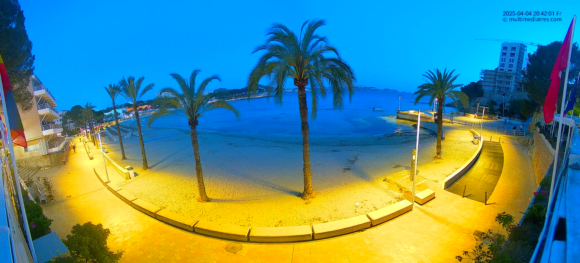 A sandy beach at twilight is illuminated by artificial lights, with palm trees lining a paved walkway next to calm blue water.