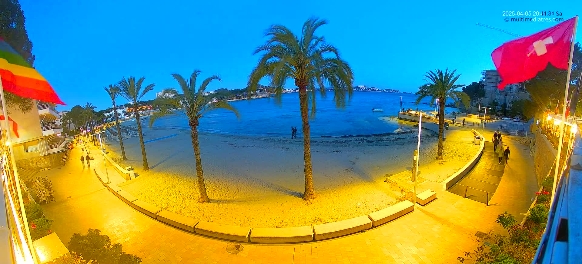 A sandy beach at twilight is bordered by a paved walkway lined with palm trees and buildings, with a few people strolling along the path.