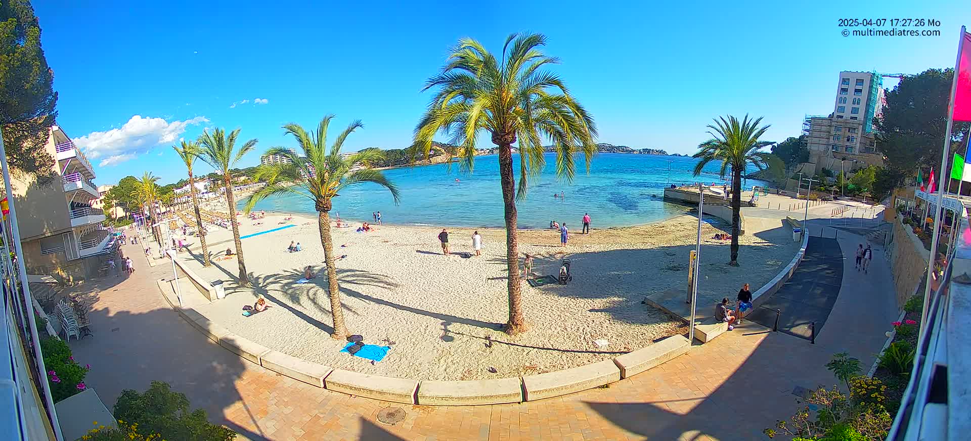 A sunny beach with calm, clear turquoise water is lined with palm trees, bordered by a paved walkway and buildings.