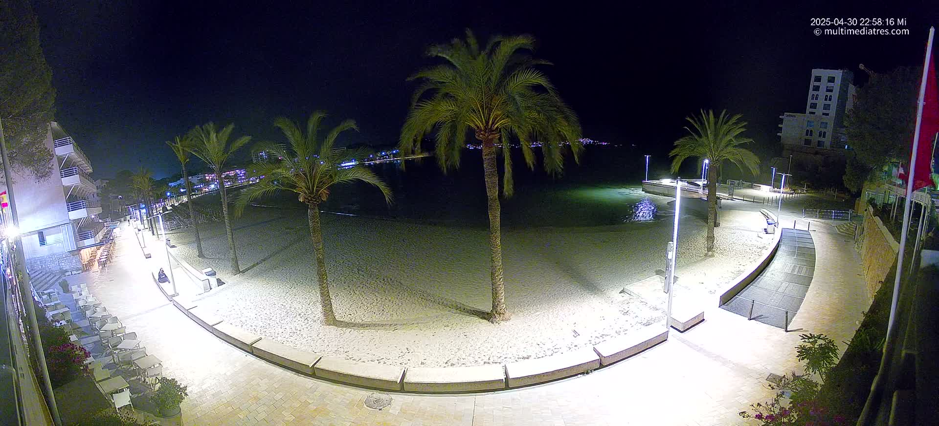 An empty, sandy beach at night is illuminated by streetlights, bordered by palm trees, buildings, and a paved walkway.