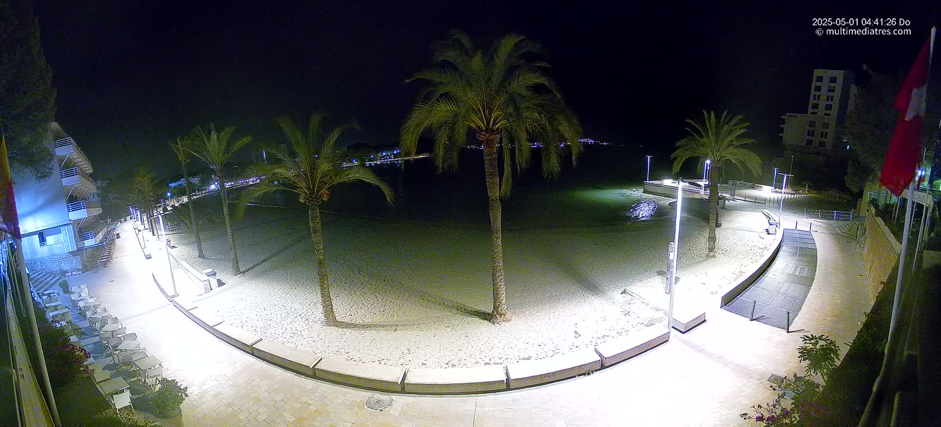 A sandy beach at night is illuminated by artificial lights, bordered by palm trees, a paved walkway, and buildings.