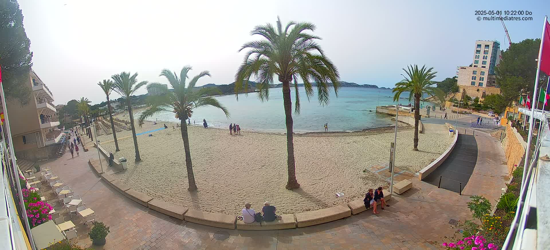 A sandy beach with calm, light blue water, palm trees, and a few people is seen from an elevated viewpoint on a partly cloudy day.