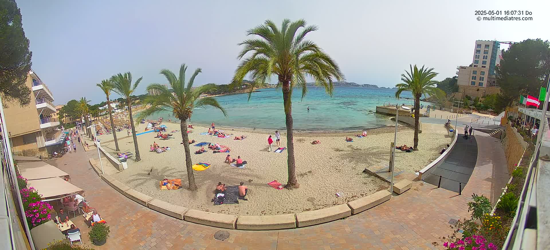 A sandy beach with scattered sunbathers is bordered by palm trees, buildings, and a paved walkway, all under a sunny sky.