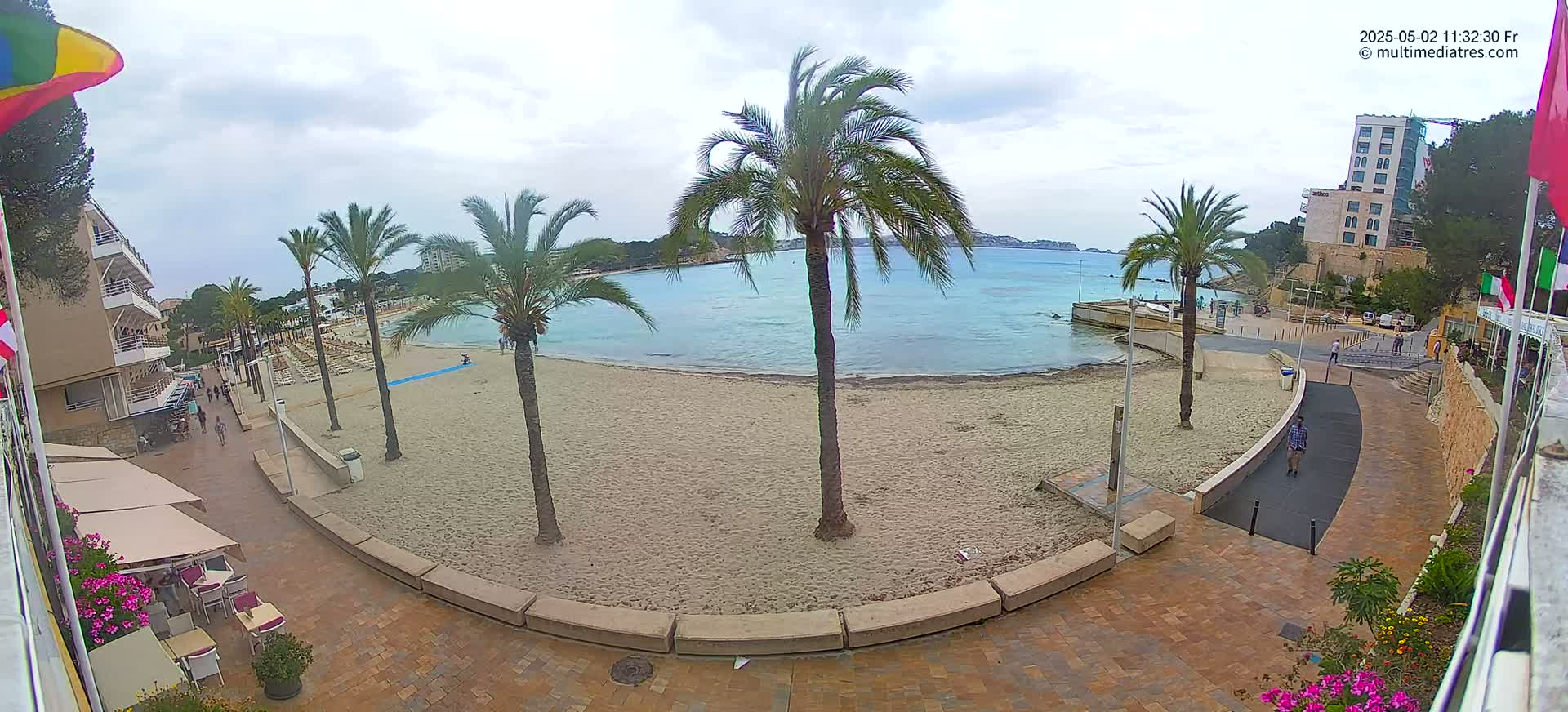 A sandy beach with palm trees and calm, light blue water is visible under a partly cloudy sky, next to a paved walkway and buildings.