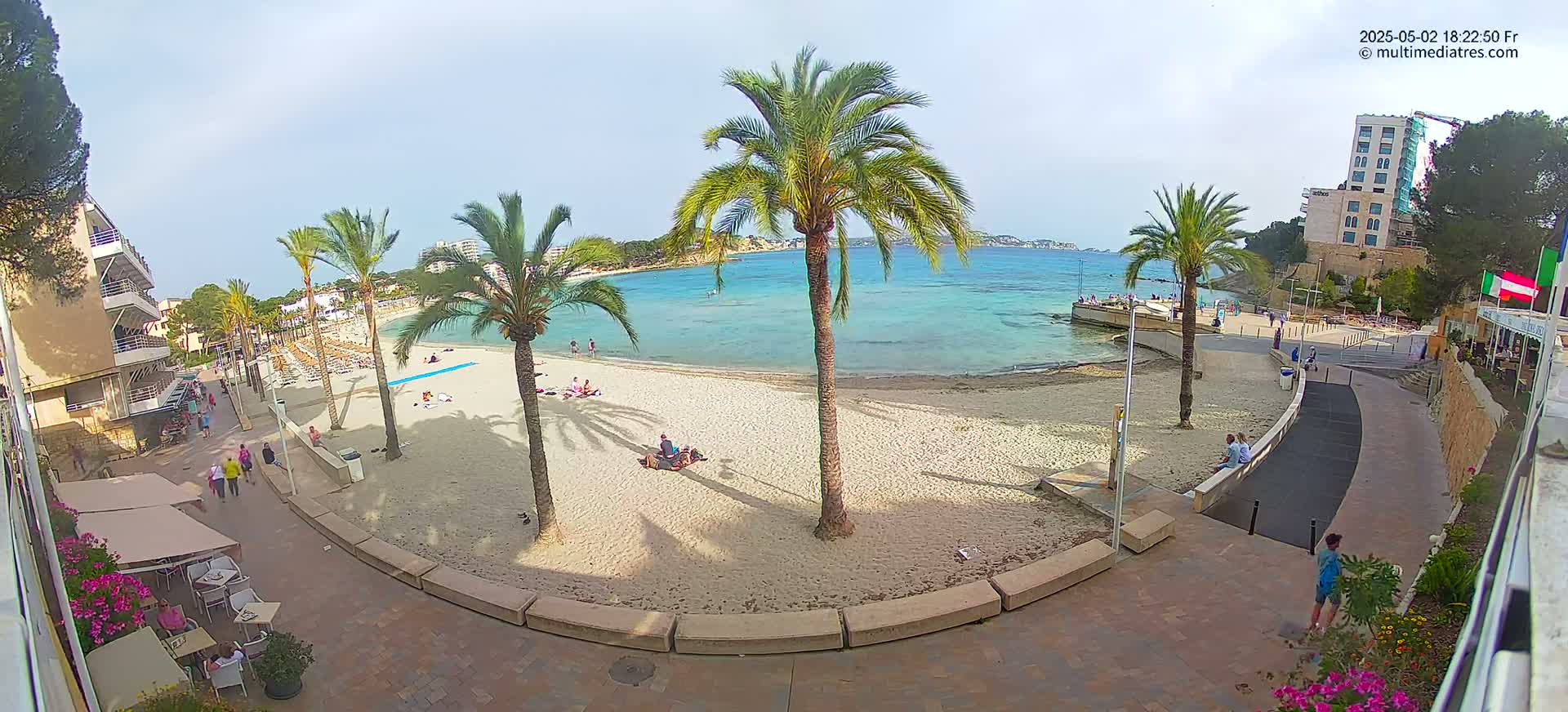 A sandy beach with palm trees and turquoise water is partially surrounded by buildings under a mostly sunny sky.