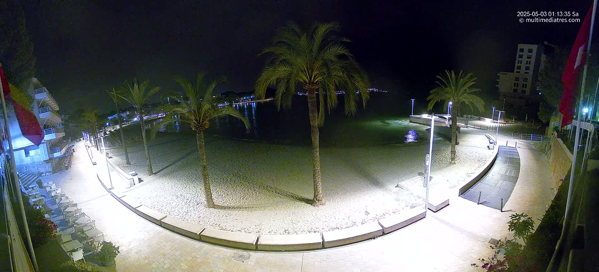 A nighttime, overhead view of a sandy beach bordered by a paved walkway, palm trees, and buildings, lit by artificial lights.