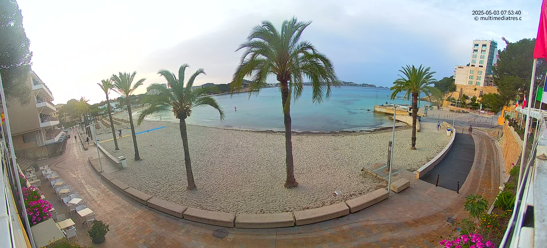 A sandy beach with calm, light blue water is visible from a beachfront promenade lined with palm trees and outdoor seating areas, under an overcast sky.
