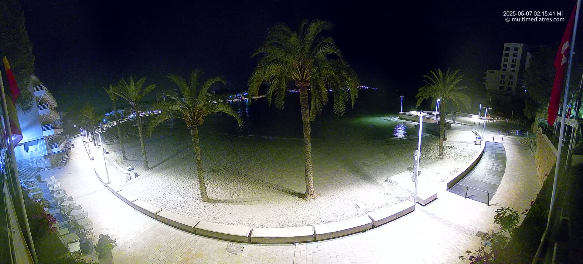A nighttime view of a sandy beach, palm trees, and a paved walkway illuminated by lights, with calm water visible in the background.