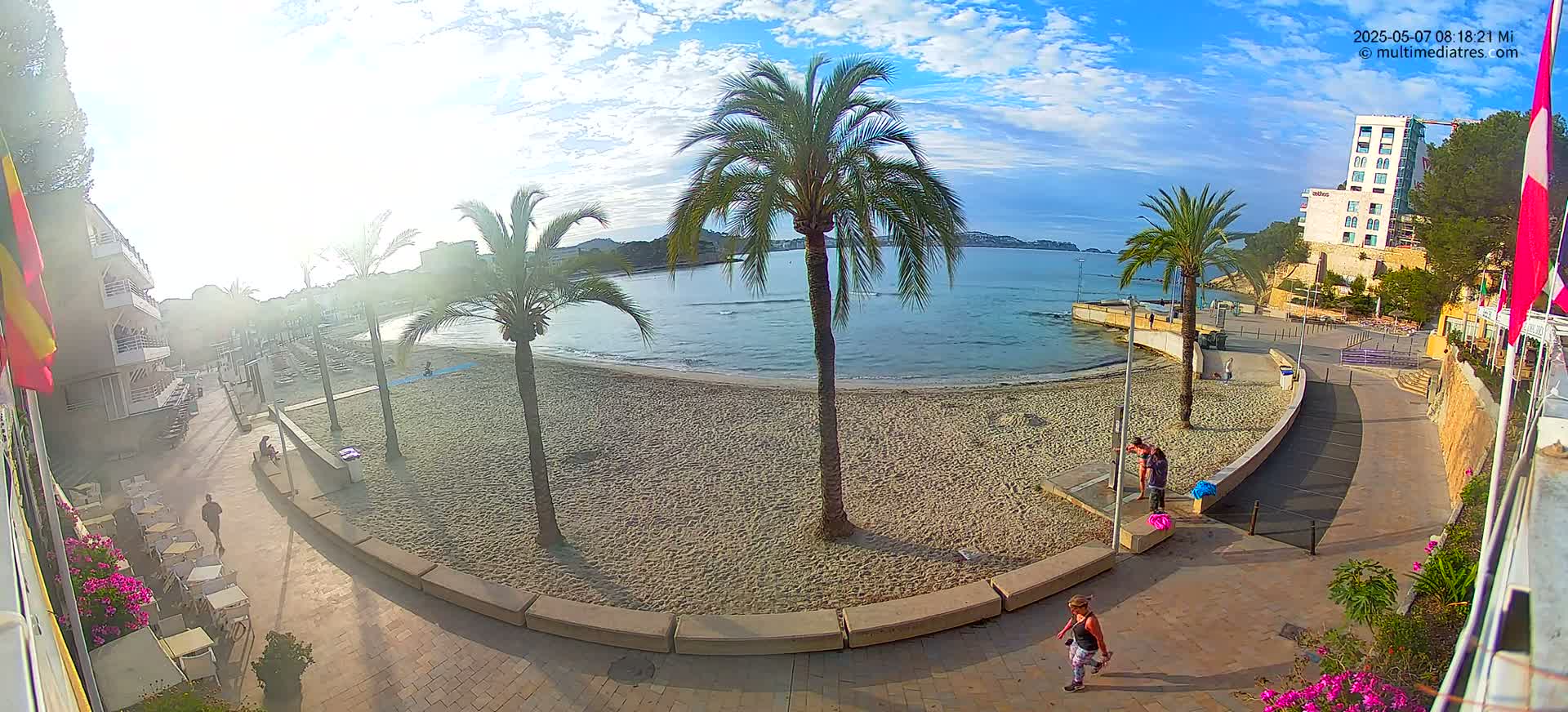 A sandy beach with palm trees and a few people is seen from above on a sunny day with some clouds.