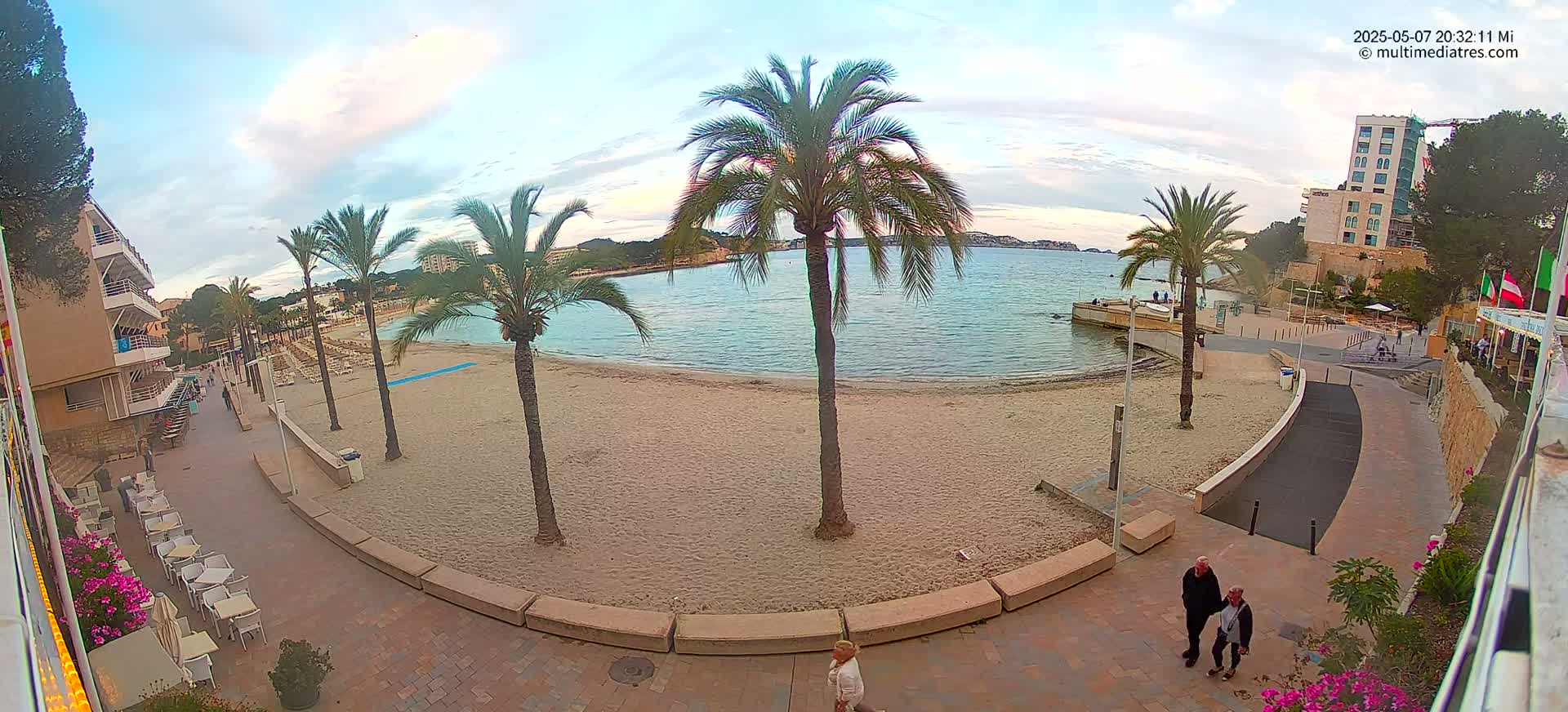 A sandy beach with palm trees, a calm bay, and a paved walkway leading to buildings under a partly cloudy evening sky.