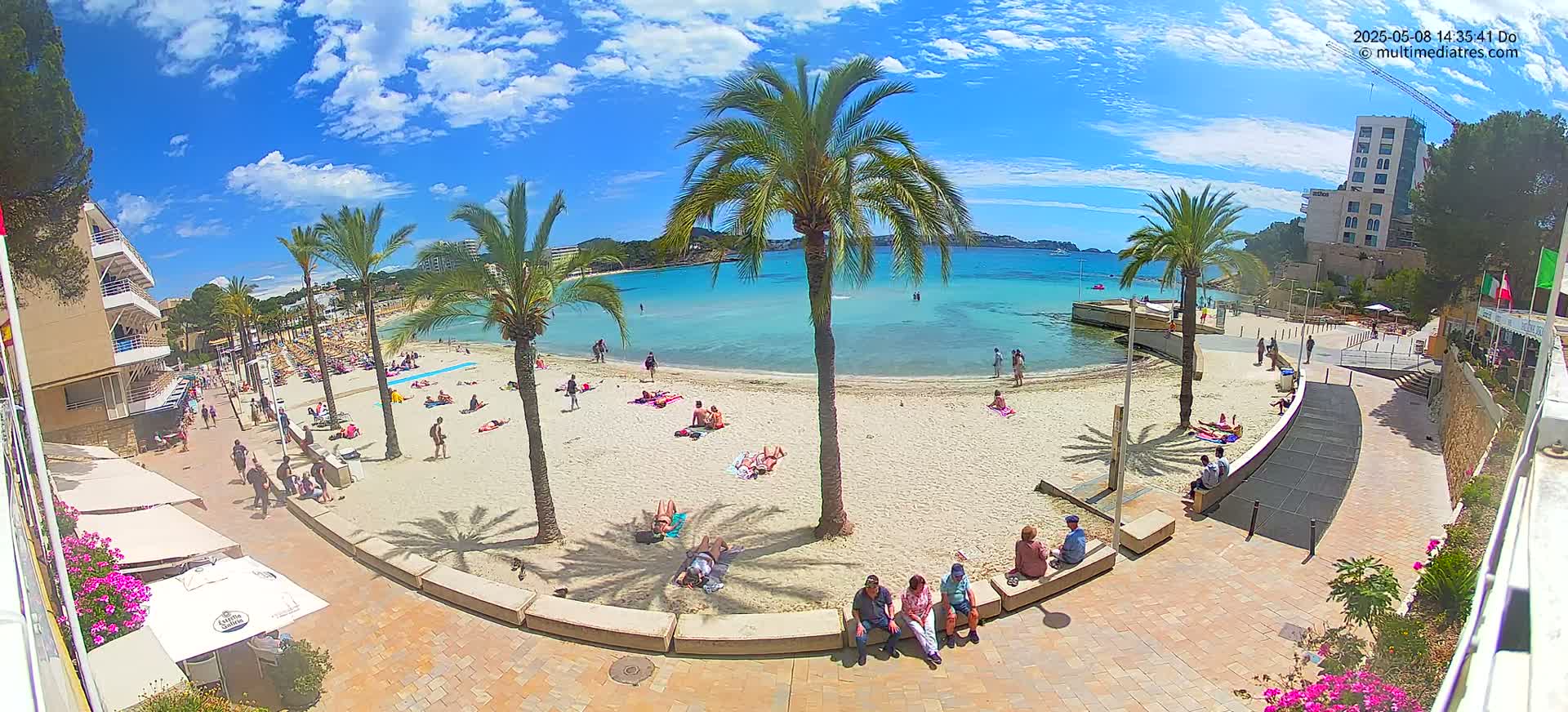 A sunny beach with clear turquoise water, palm trees, and many people relaxing on the sand and in the water.