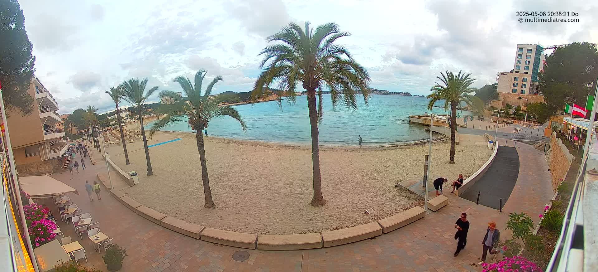 A sandy beach with calm, light blue water is bordered by a paved walkway lined with palm trees, buildings, and people.