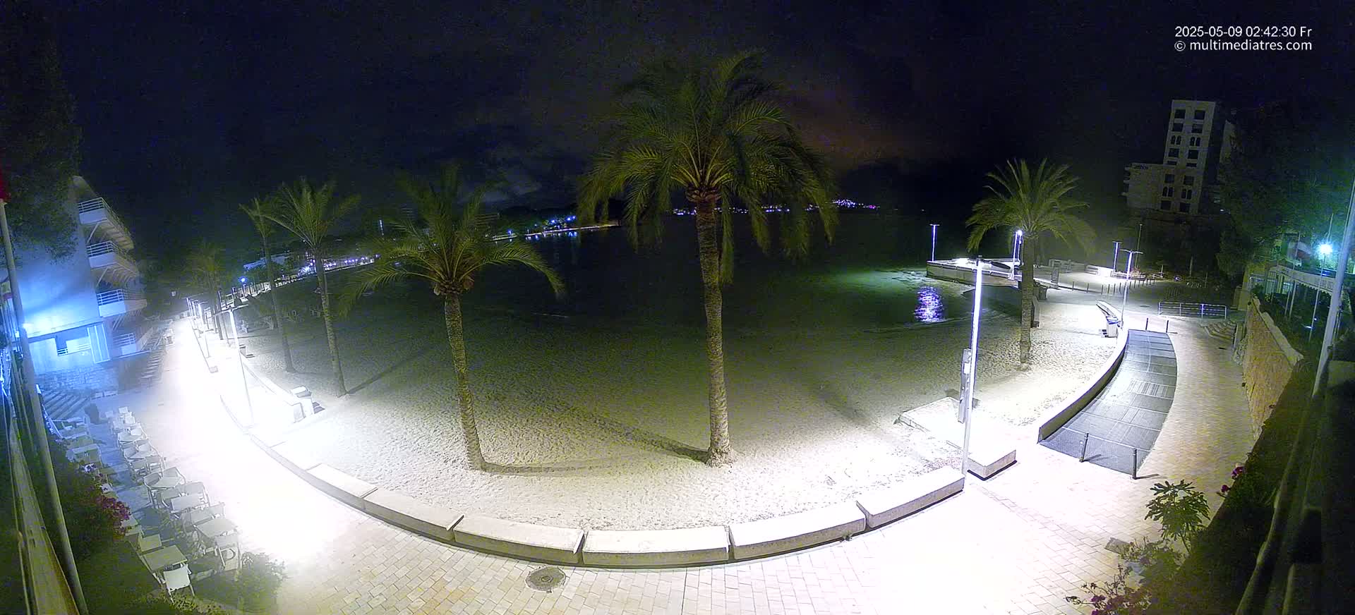 A nighttime view of a sandy beach with palm trees, illuminated walkways, and buildings in the background under a clear, dark sky.
