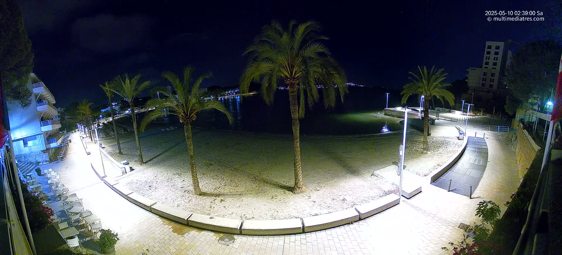 A nighttime view of a sandy beach with palm trees, a paved walkway lined with outdoor seating, and buildings visible in the background under a partly cloudy sky.