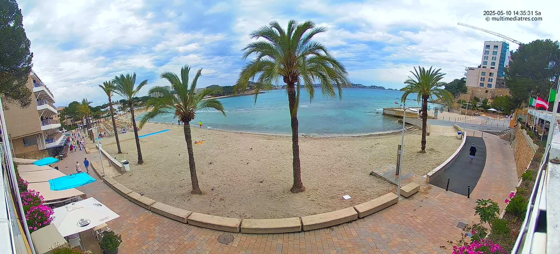 A sandy beach with palm trees and several people on it, under a partly cloudy sky, is bordered by buildings and a paved walkway.