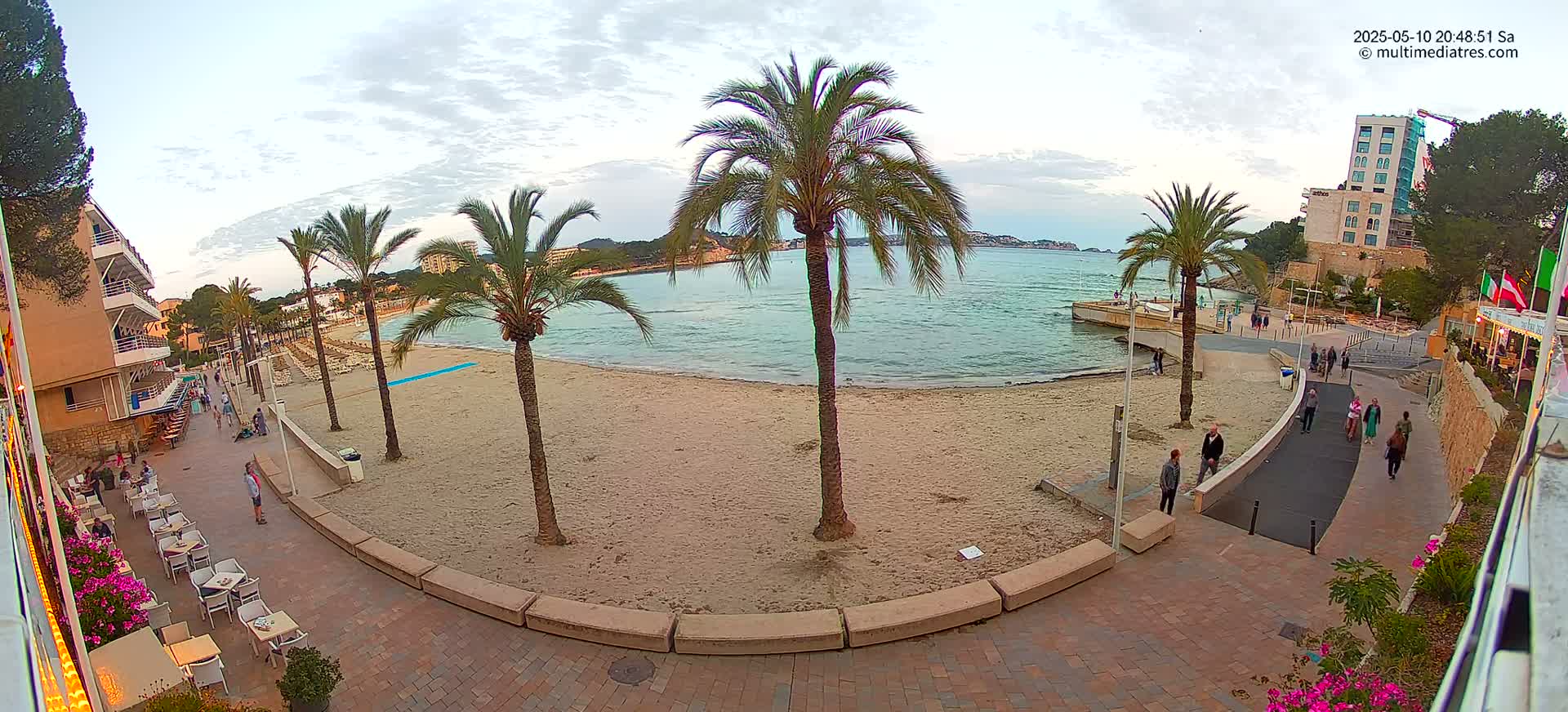 A sandy beach with palm trees and several people walking along a paved walkway next to an outdoor restaurant, under a partly cloudy sky.