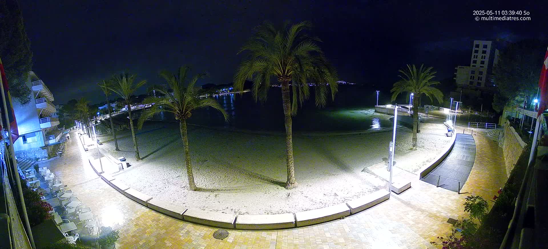 A nighttime view of a sandy beach, palm trees, and a paved walkway illuminated by lights, overlooking calm water.