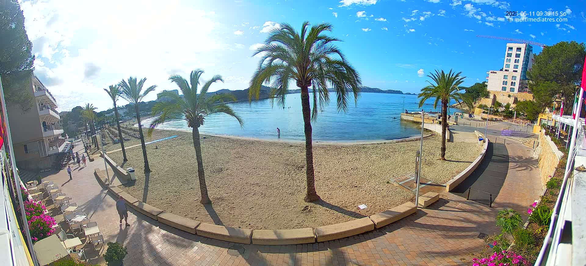 A sandy beach with palm trees and several people is visible from an elevated viewpoint on a sunny day with a few clouds.