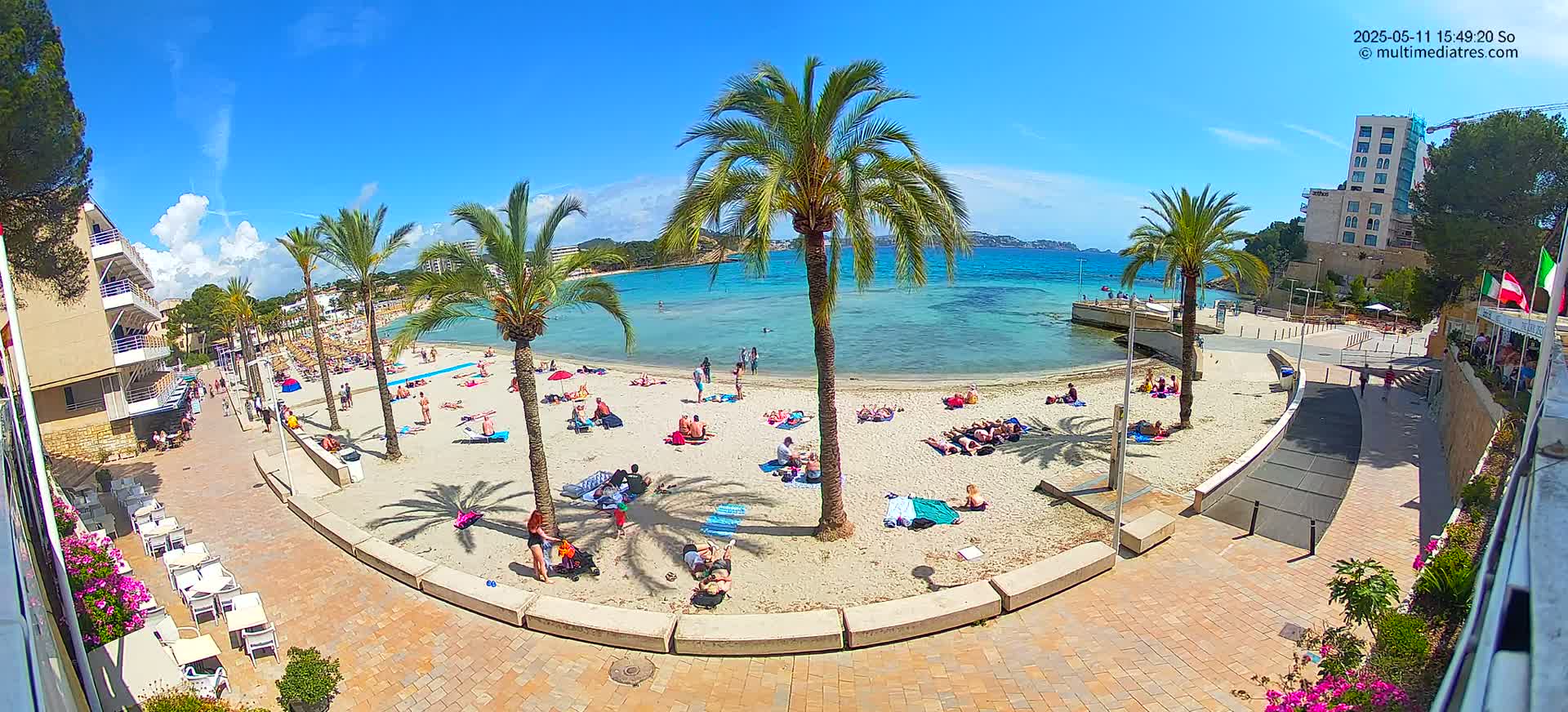 A sunny beach scene shows people relaxing on a sandy beach, next to calm, clear blue water, under a mostly clear blue sky, surrounded by palm trees and buildings.