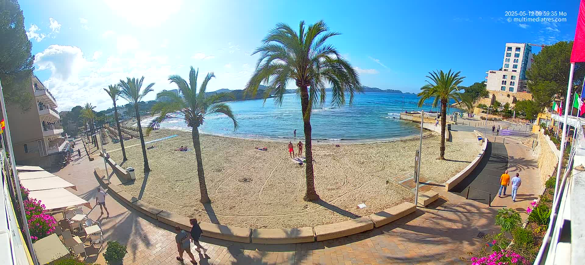 A sandy beach with palm trees and a few people, situated next to a paved walkway and buildings, under a bright, sunny sky.