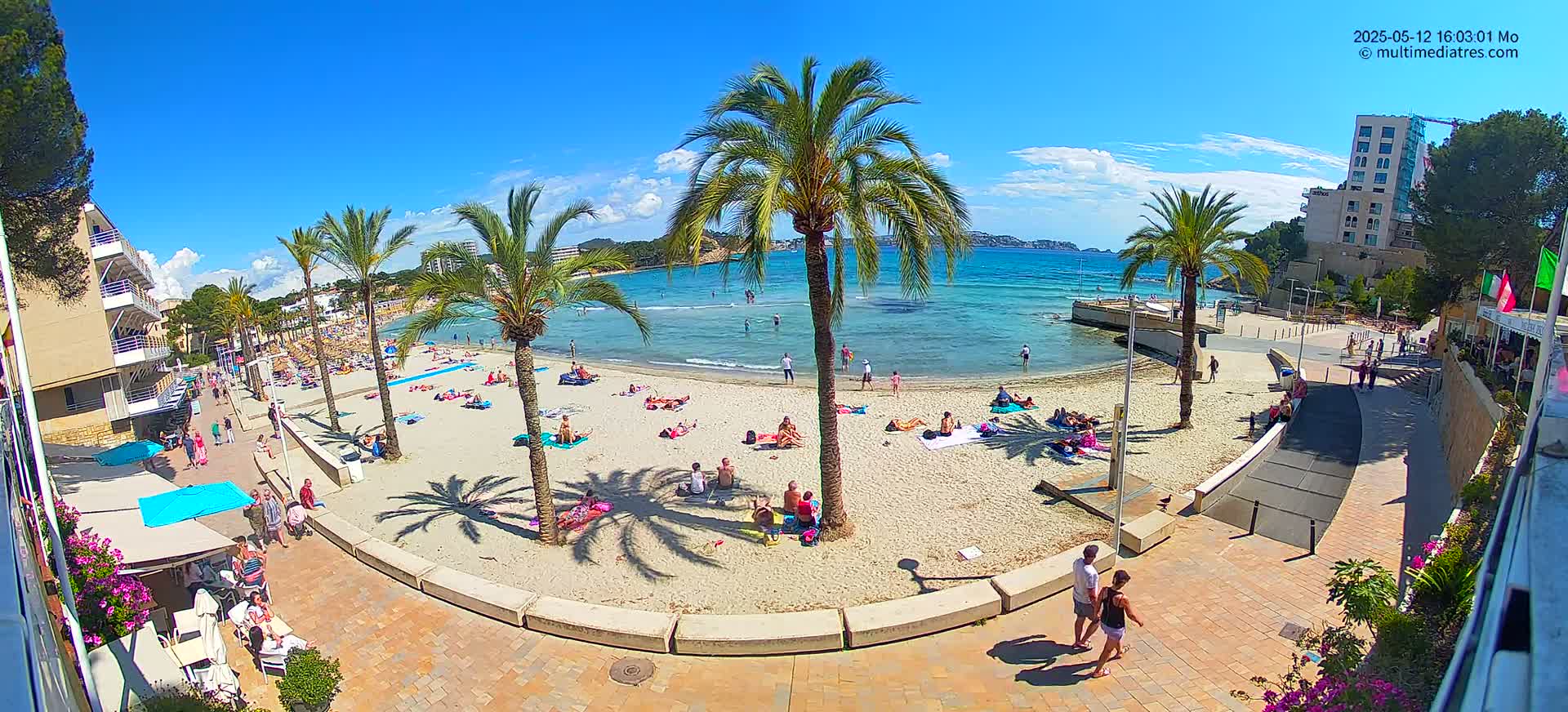 A sunny beach scene shows people relaxing on a sandy beach near calm, blue water under a mostly clear sky, with palm trees and buildings lining the shore.