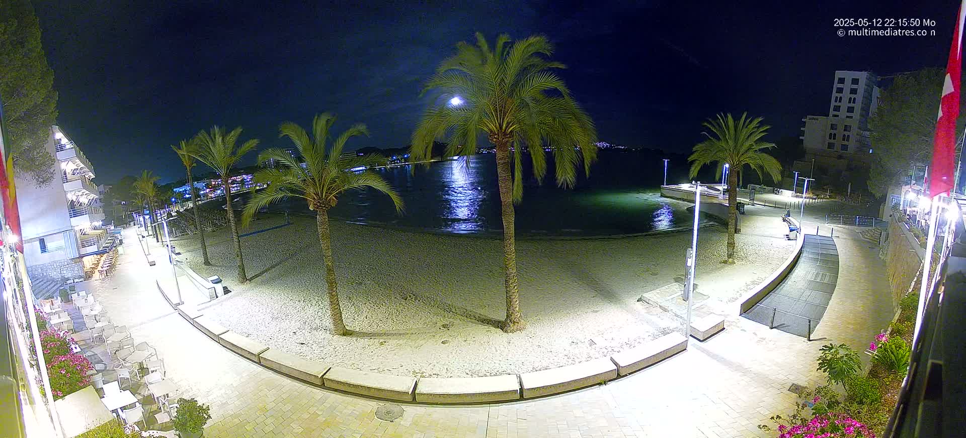 A moonlit night scene shows a sandy beach with palm trees, a paved walkway, and buildings, all illuminated by artificial lights.