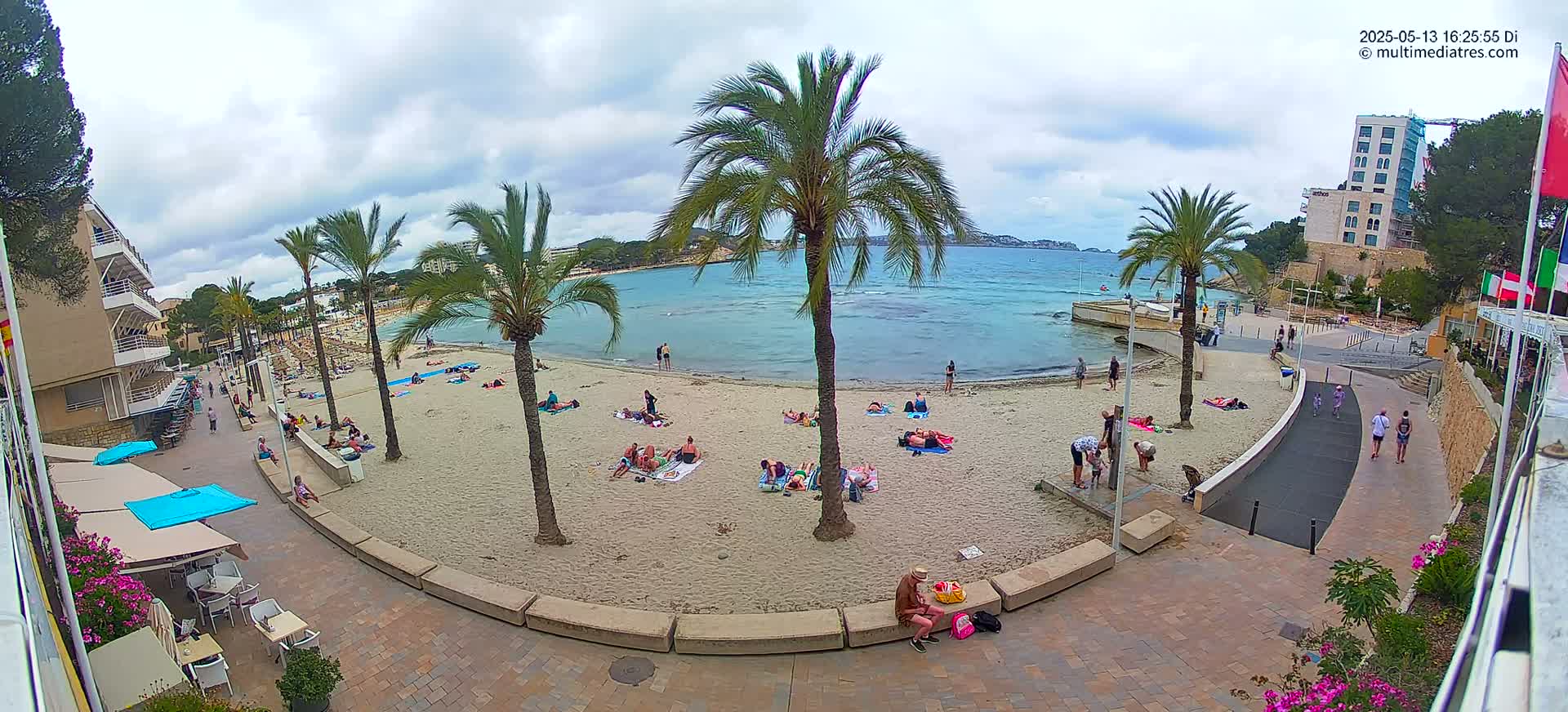 A sandy beach with people sunbathing and swimming, palm trees, and buildings, under a partly cloudy sky.