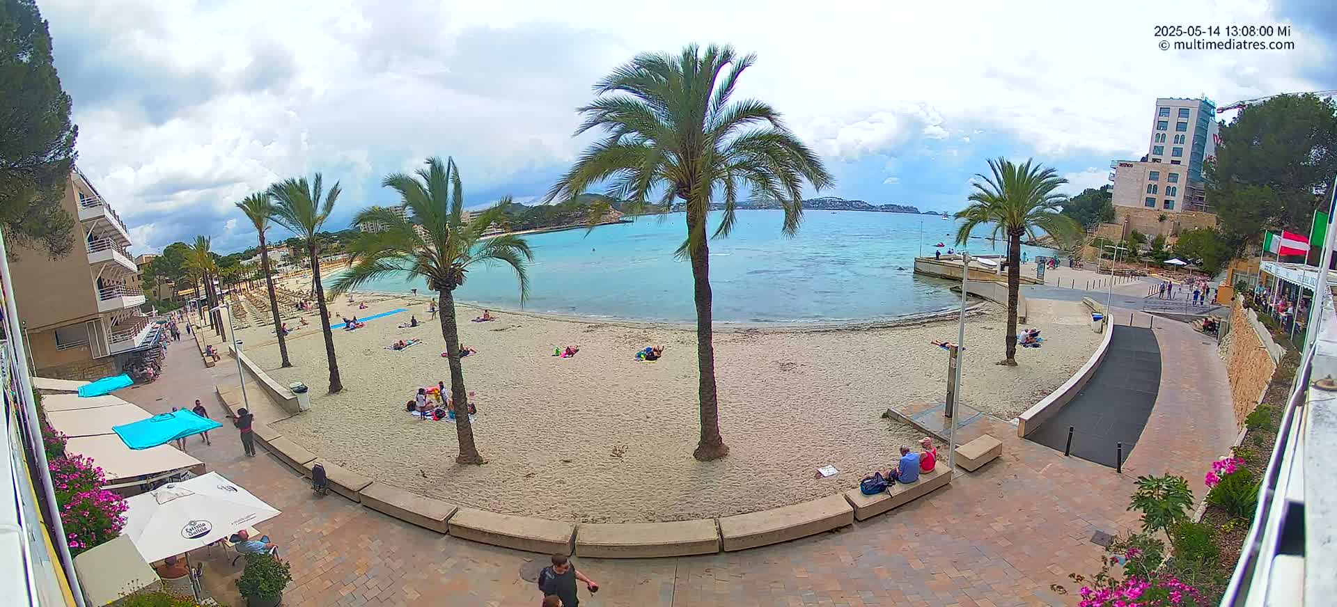 A partly cloudy day at a sandy beach with palm trees, several people sunbathing and relaxing, and buildings along the shore.