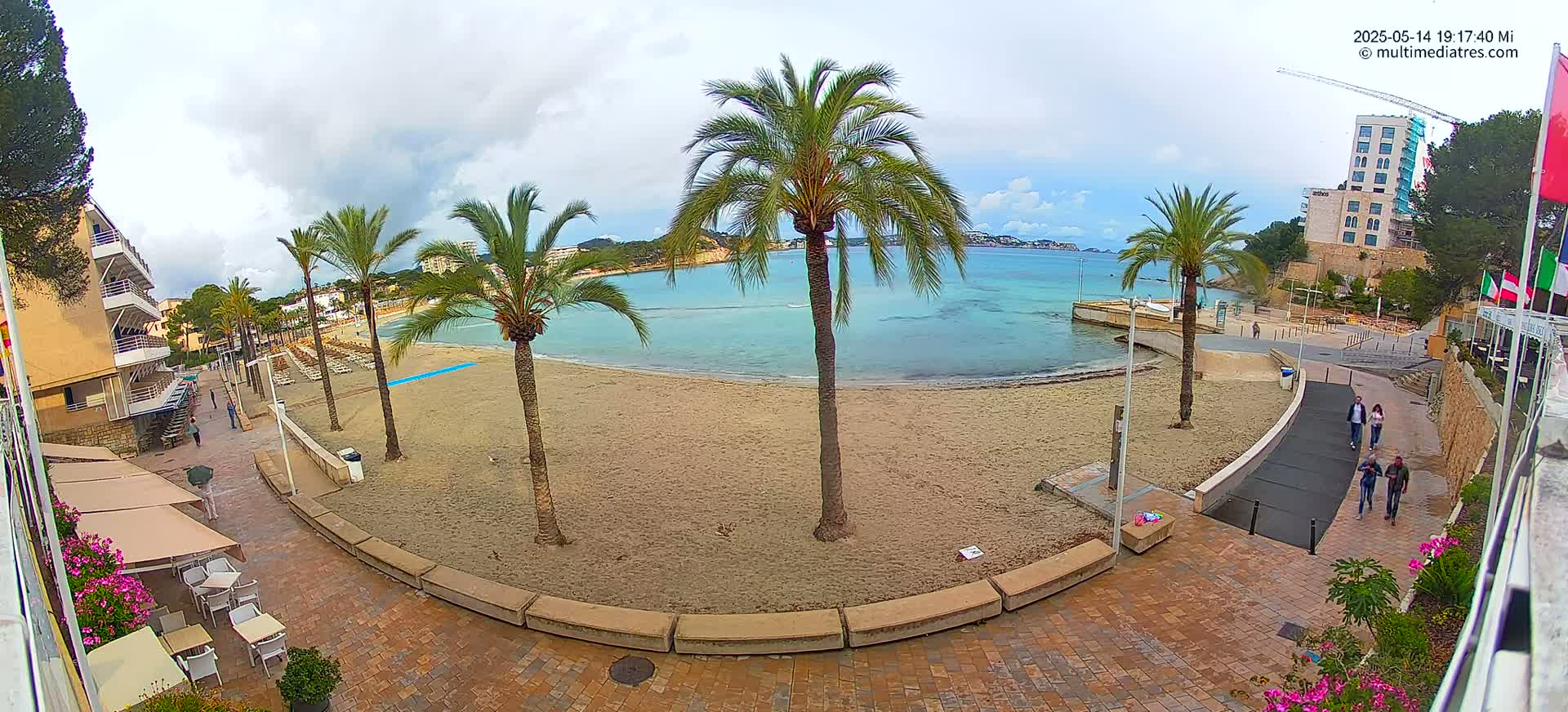 A sandy beach with palm trees, buildings, and a few people walking along a paved walkway on an overcast day.