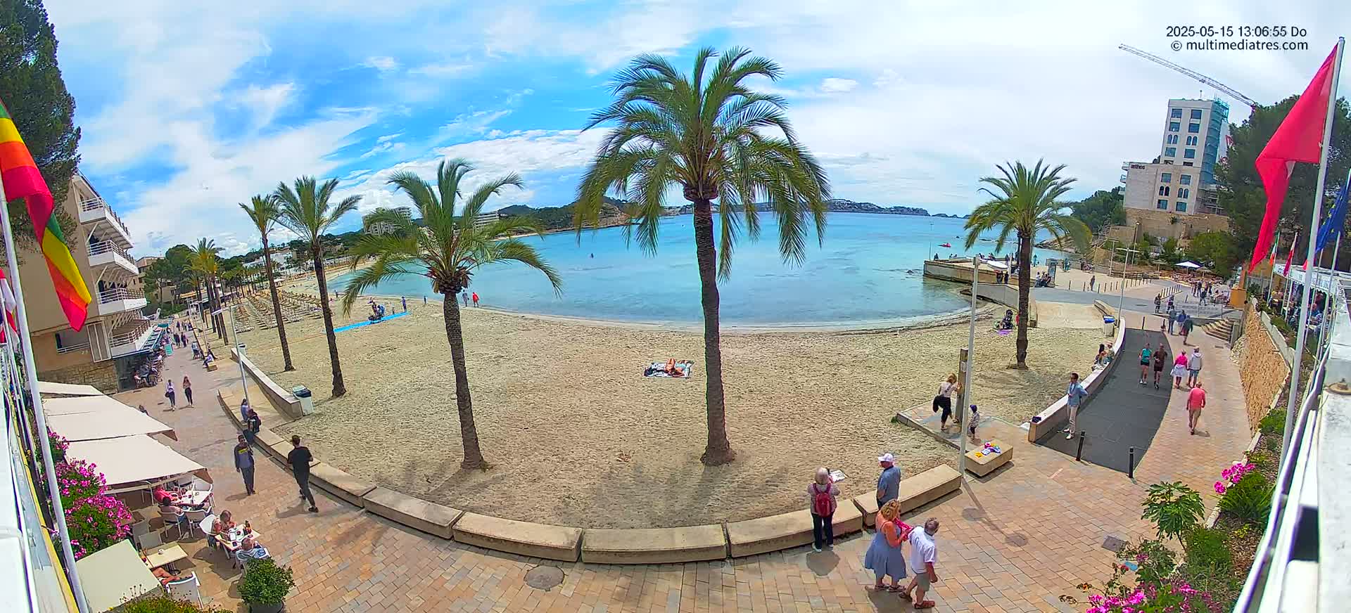 A sandy beach with calm, blue water is bordered by a paved walkway lined with palm trees, buildings, and people under a partly cloudy sky.