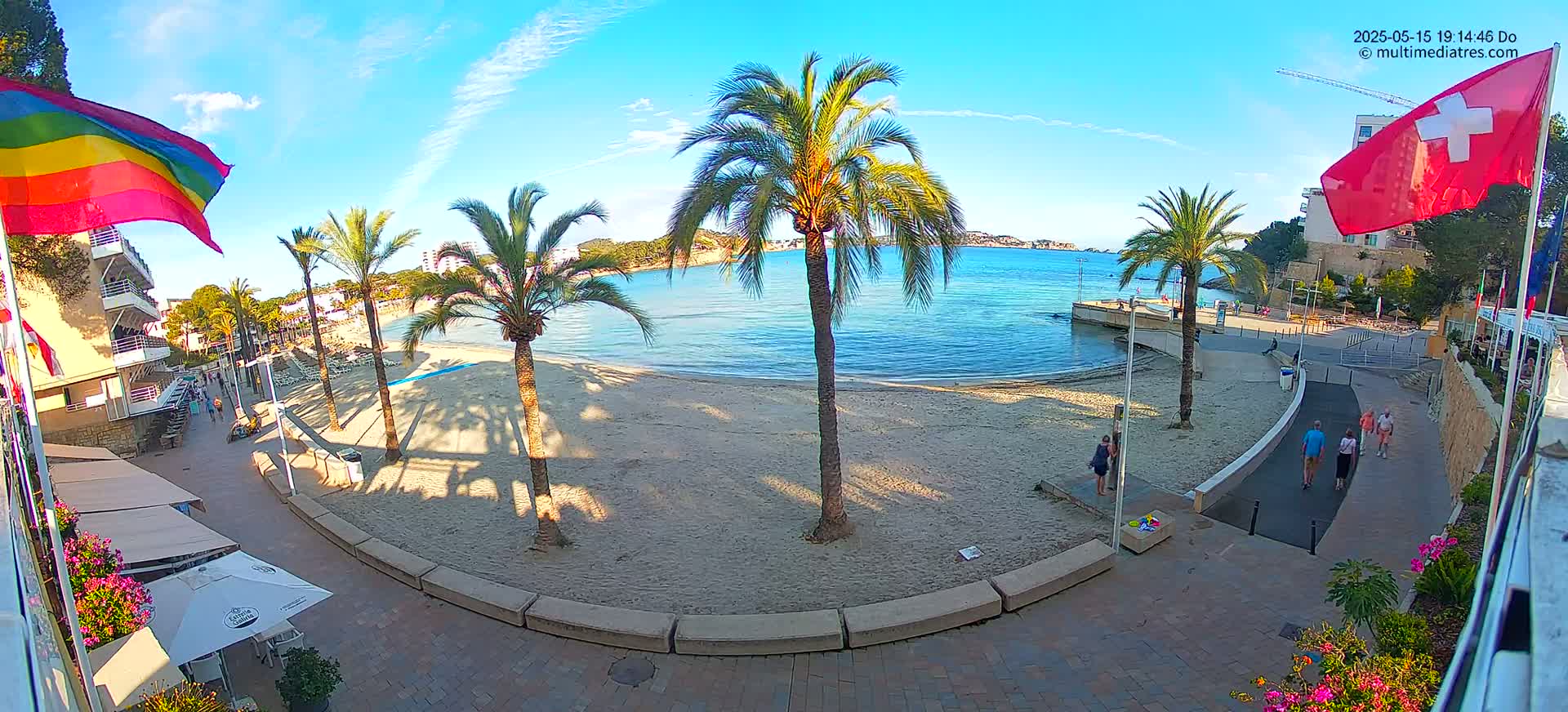 A sandy beach with palm trees and calm blue water is viewed from a seaside promenade on a sunny day.