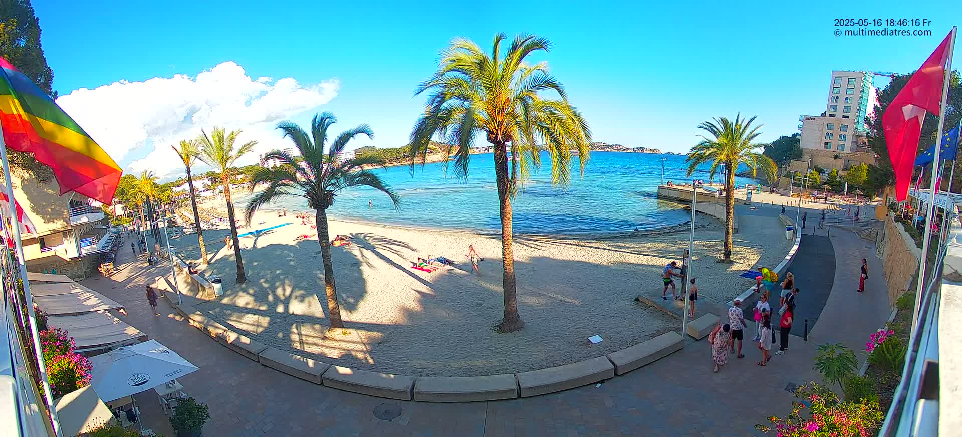 A sunny beach with clear blue water, palm trees, and people relaxing on the sand and walking along a paved promenade.