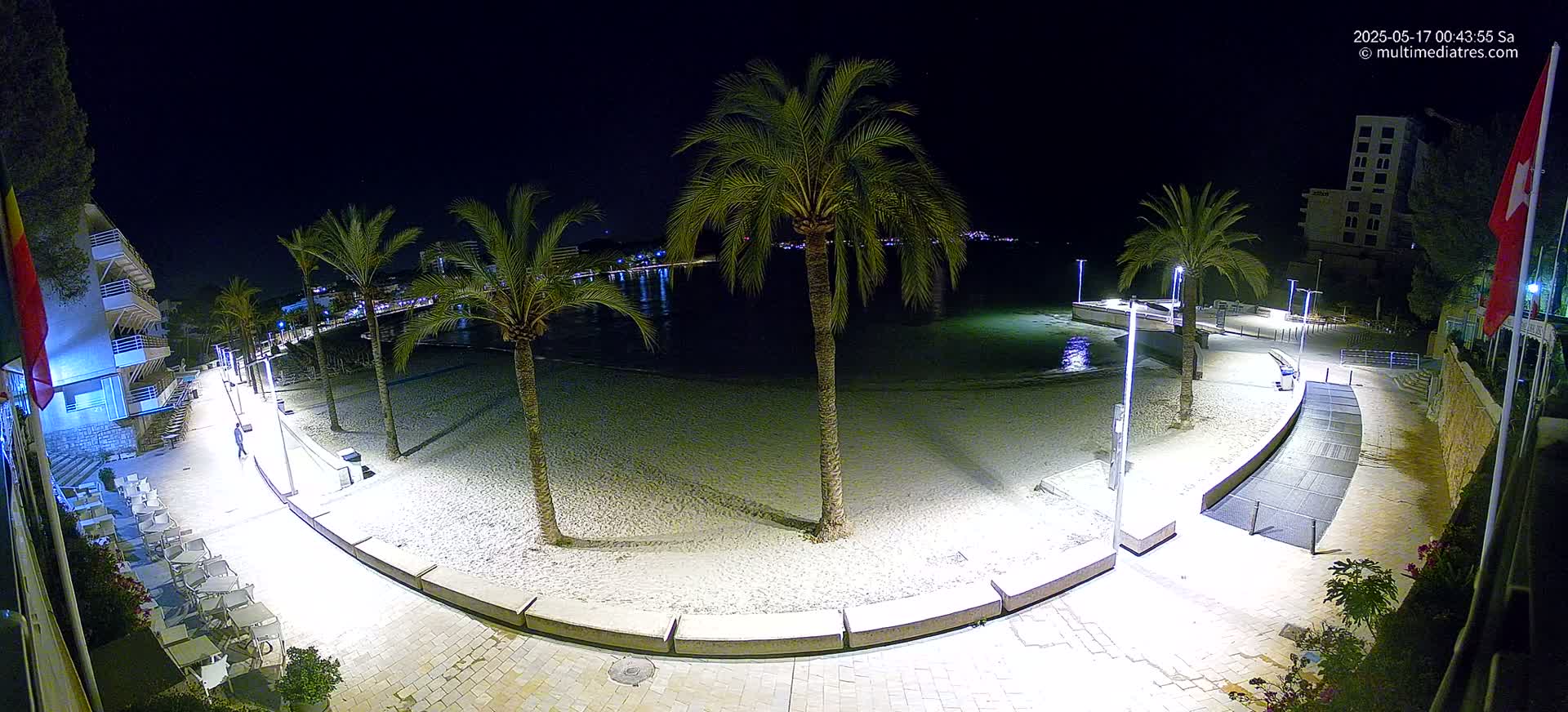 A nighttime, illuminated view of a sandy beach bordered by a paved walkway, palm trees, and buildings, with calm water visible beyond.