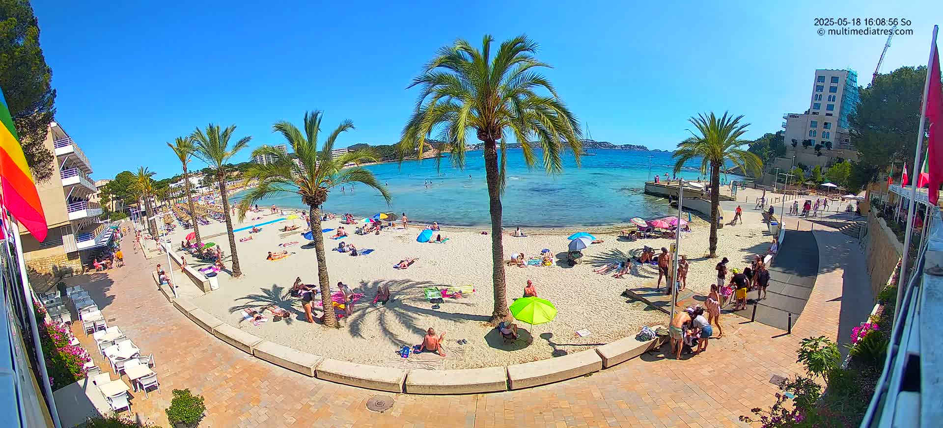 A sunny beach scene shows numerous people relaxing on the sand under umbrellas, with palm trees lining a paved walkway leading to buildings and a calm, turquoise bay.