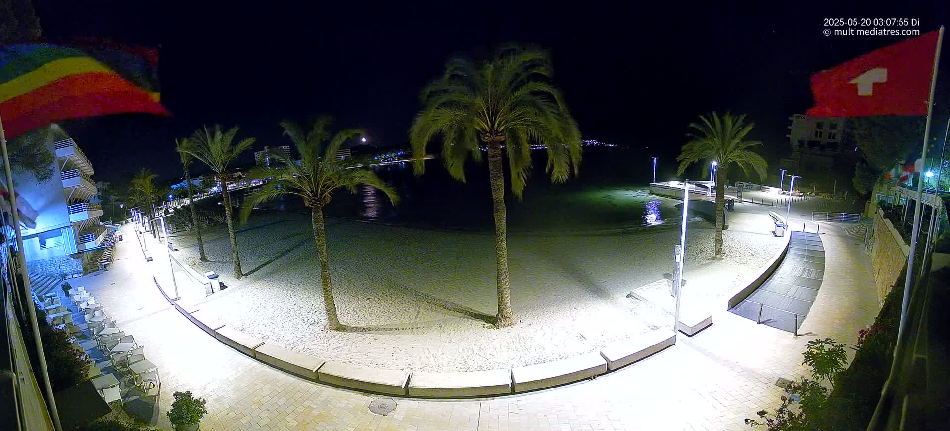 A nighttime view of a sandy beach illuminated by lights, bordered by a paved walkway, palm trees, and buildings.